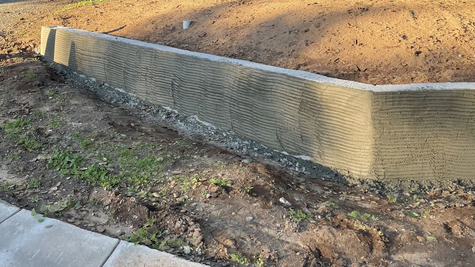 Long retaining wall curving around a graded bed beside a new concrete walk